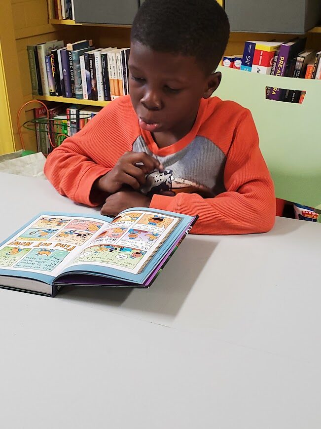 A Black male child wearing an orange and gray shirt sitting at a white table reading a book open flat in front of him