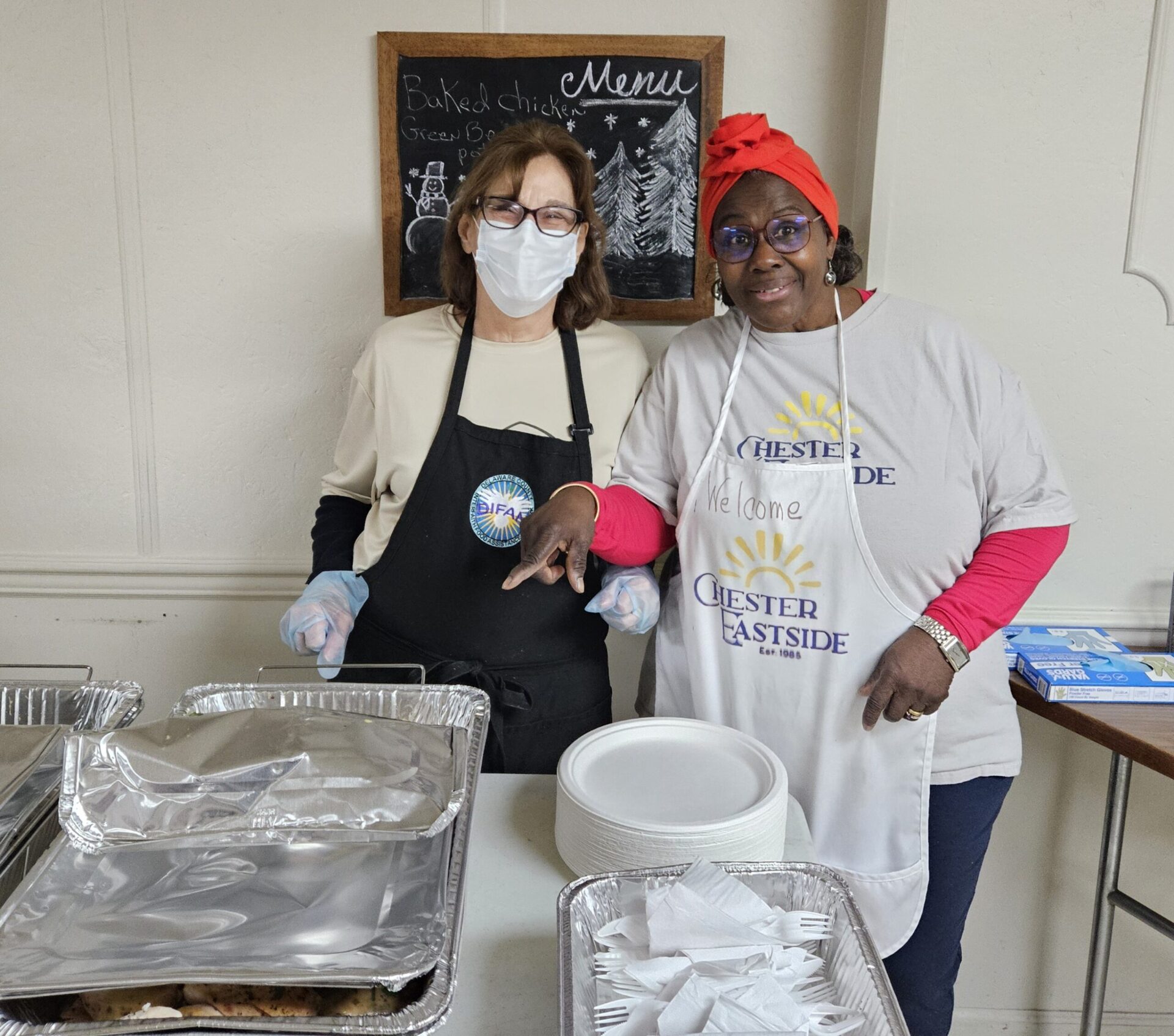 Two women wearing aprons, one Black, one white and wearing a medical mask, are both pointing at a foil covered pan of food on a table.