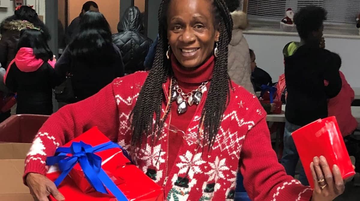 A Black woman with long braids is wearing a red and white sweater. She is cradling a wrapped present in her right arm and holding a red object in her left hand.