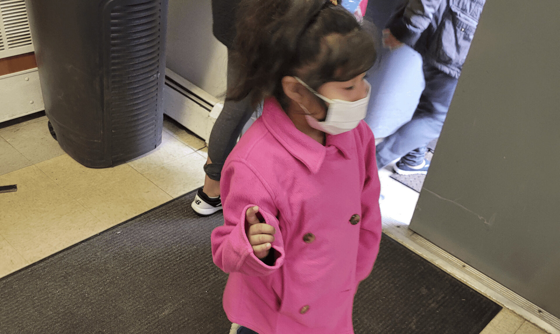 A little girl wearing a white medical mask and a pink coat