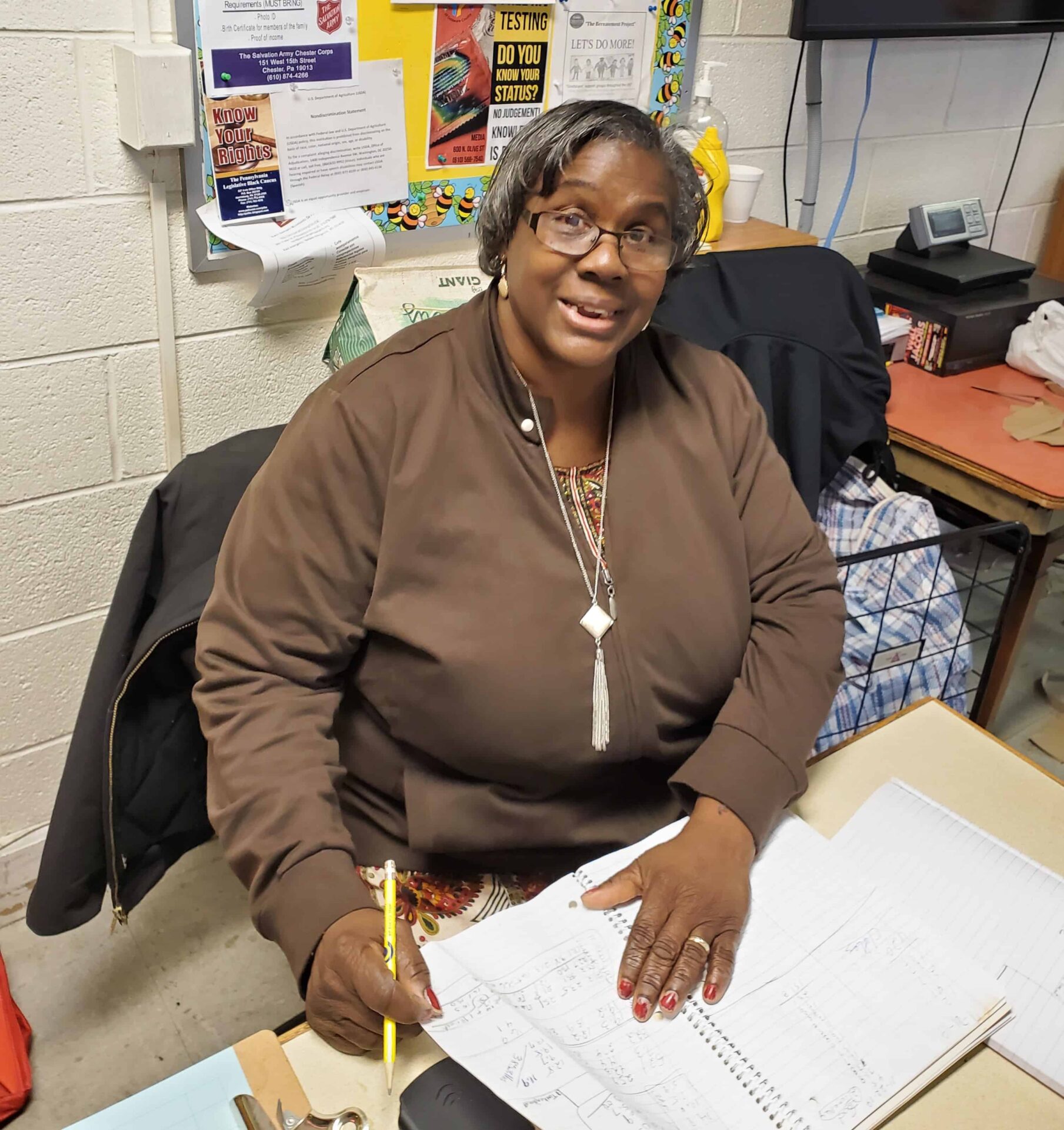 A Black woman with gray hair wearing a zipped up brown jacket. She is sitting at a desk with a notebook open and a pencil in her hand