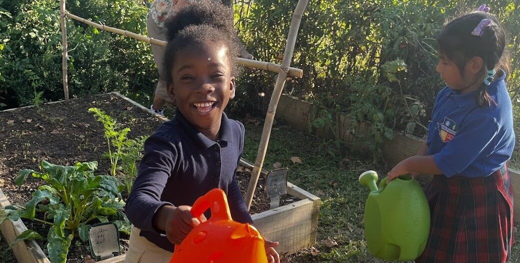 A smiling young girl holds an orange watering can. Another girl is to her left holding a green watering can.