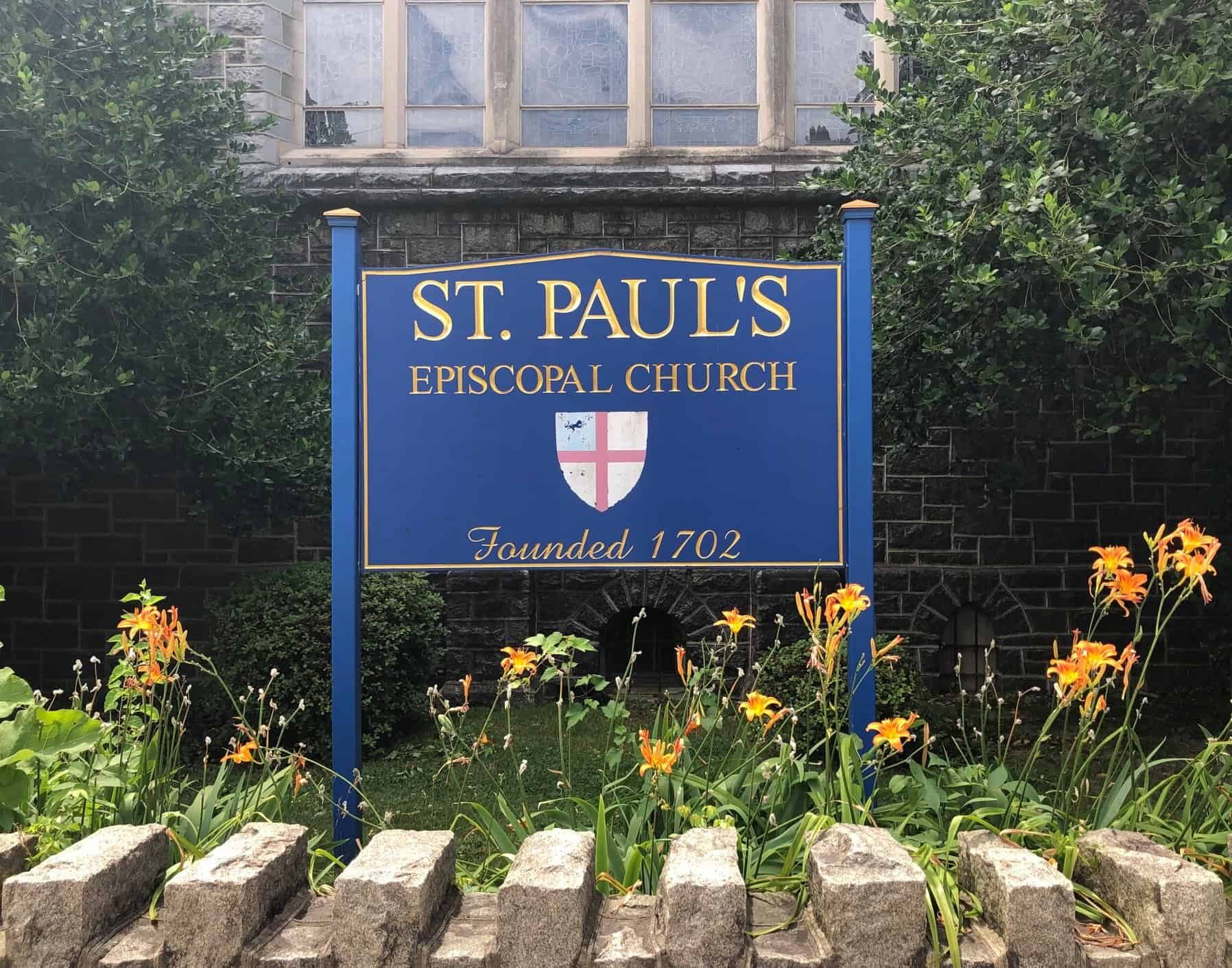 A blue wooden sign in a garden behind a small stone fence. The sign says St Paul's Episcopal Church, Founded 1702