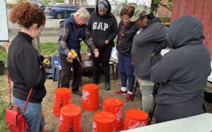 An older white man holding a drill is speaking to a group of five people. They are standing in a circle around upturned orange Home Depot buckets.