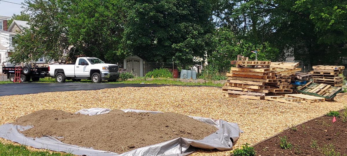 A pile of dirt on a lot covered with wood shavings. There are pallets in the rear and a white truck parked to the left.