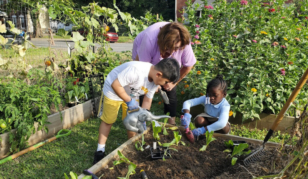 A woman is bending over supervising two children one bending and watering, one squatting and digging, in a garden