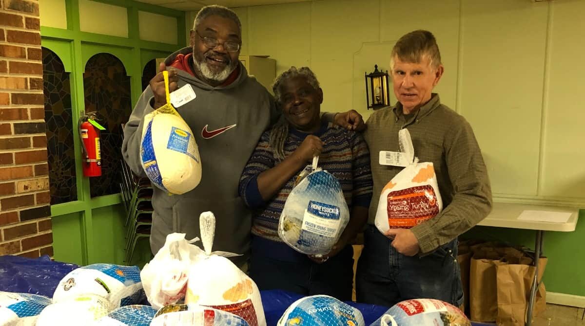 Two men holding frozen turkeys behind a table with more frozen turkeys