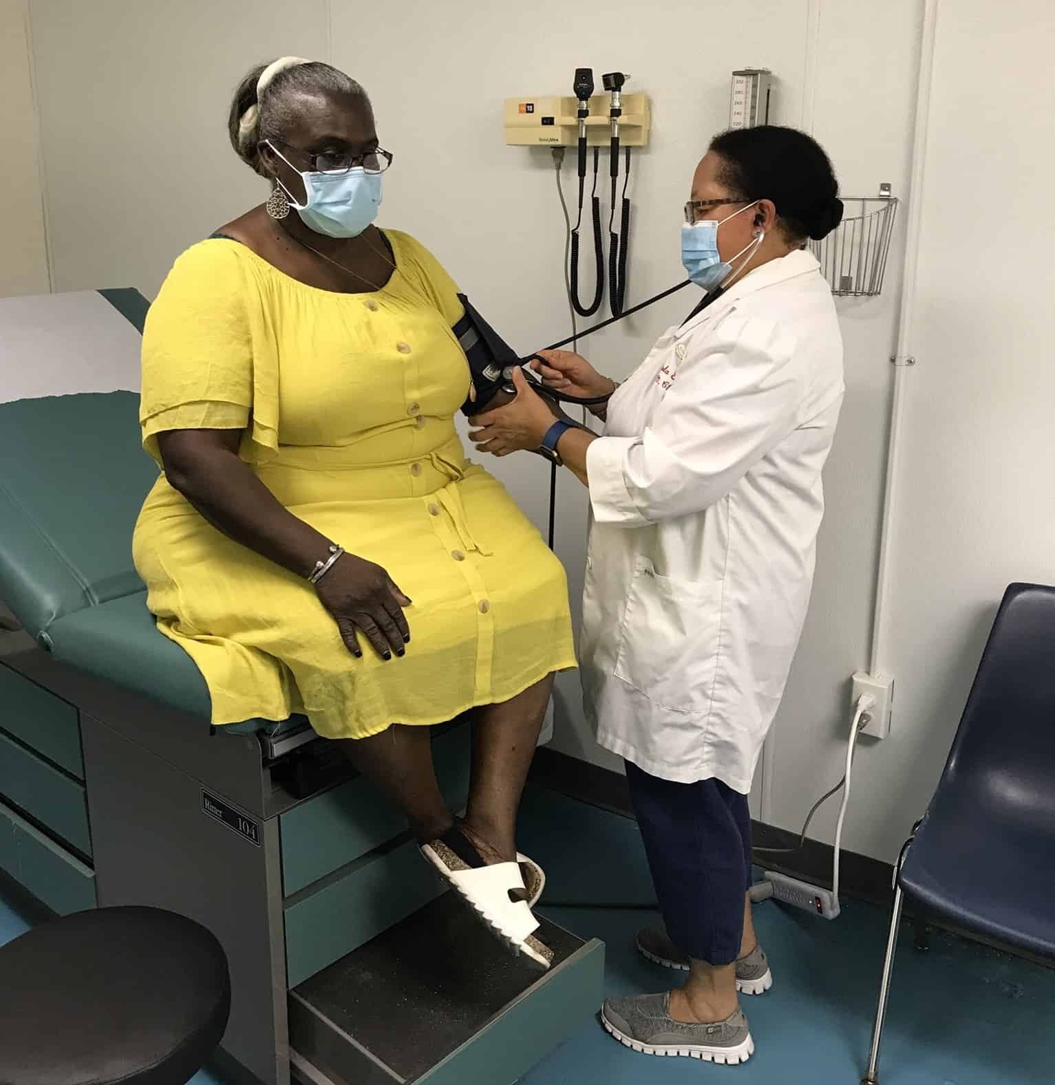 A Black female doctor wearing a medical mask and a white coat taking the blood pressure of a Black woman wearing a yellow dress, glasses and a medical mask