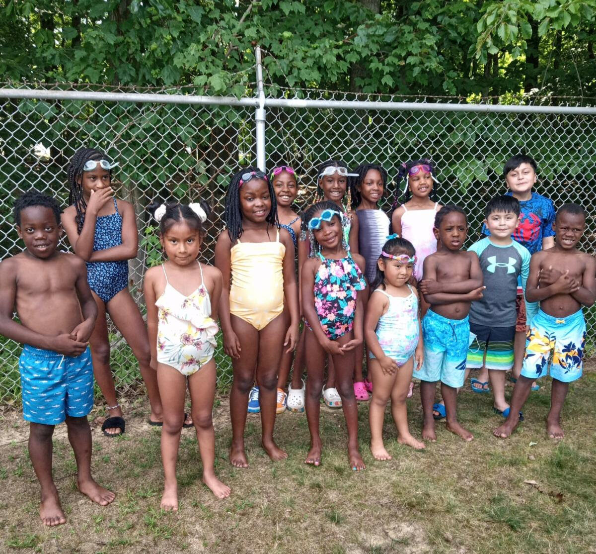 A large group of kids in swimsuits standing in front of a metal fence