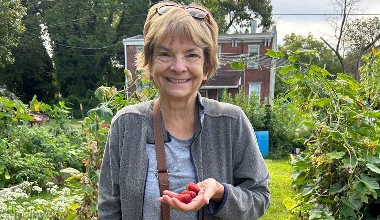 A white woman with brown hair wearing a blue cardigan, blue shirt and sweatpants. A striped bag is slung across her body. Her left hand is full of berries and she is leaning on a cane with her right.