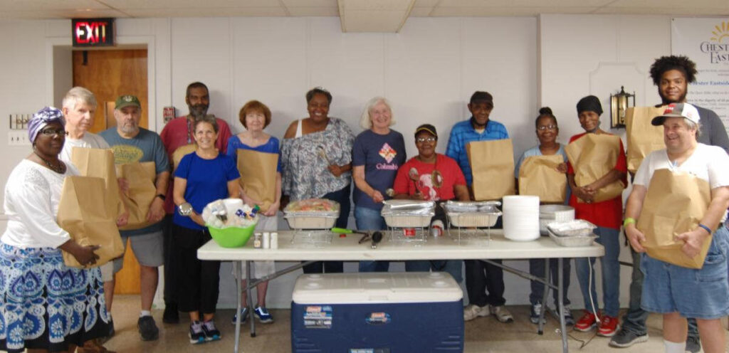 A group of volunteers standing around a table. Some are holding large filled paper bags. There are several serving dishes of food on the table.