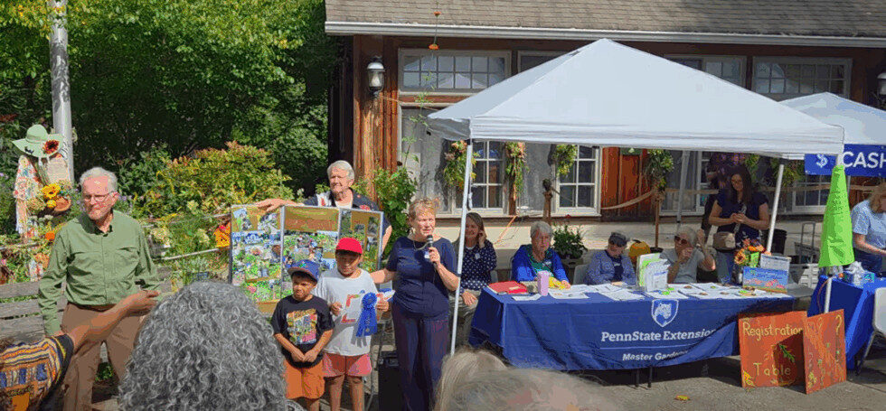 A white woman is speaking into a microphone. There are two children to her left, two men behind and to the left of her. There are several people seated at a table behind and to the right of her.