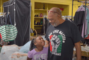 An older Black man is wearing glasses and a Black Lives Matter t-shirt. He is hugging a young Black girl. The girl is wearing a pink top with Minnie Mouse on it. They are standing in a room full of bags, clothes and shoes.